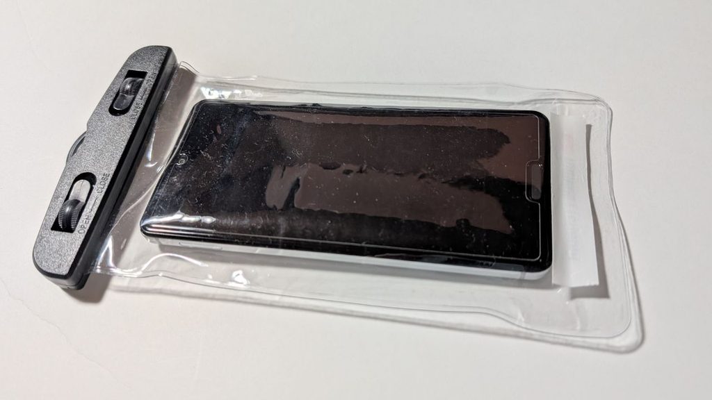 スマートフォンを入れた透明スマホポーチを上から撮影した写真。黒い留め具とポーチ全体の形が見える