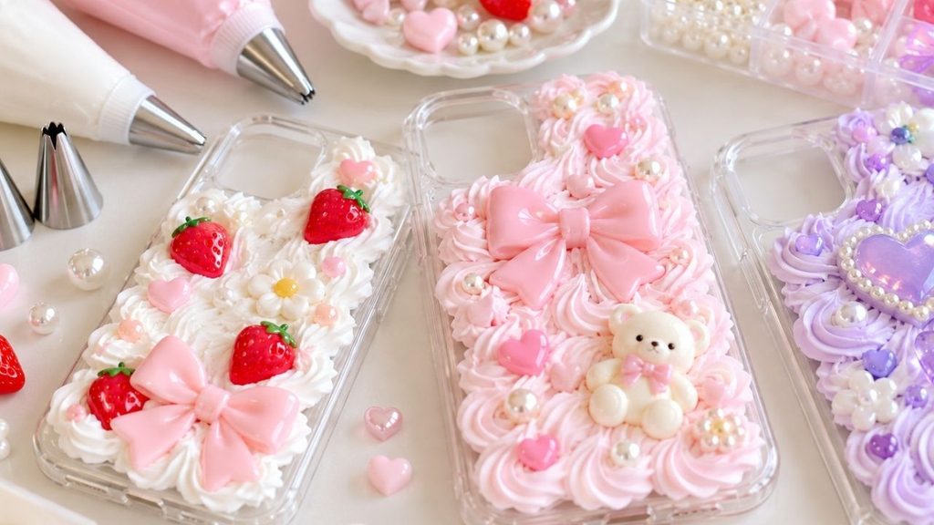 白・ピンク・紫のホイップクリーム風の飾りに、いちご、リボン、ハート、花、くまのパーツを組み合わせた透明スマホケース3点のAIイメージ画像